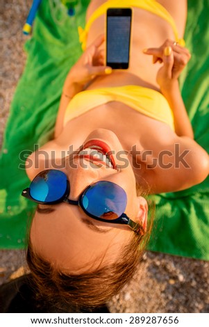 Young funny woman in swimsuit reading with mobile phone lying on the green towel on the beach. Top view focused on the face.