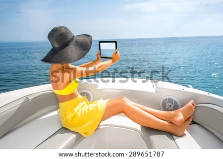 Young woman in yellow swimsuit and skirt photographing seascape with digital tablet lying on the yacht.