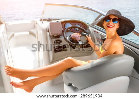 Young and pretty woman in yellow skirt and swimsuit with hat and sunglasses driving luxury yacht in the sea.