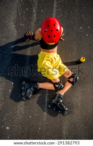Young sport woman in yellow skirt and red helmet with rollers lying on the road. Top view