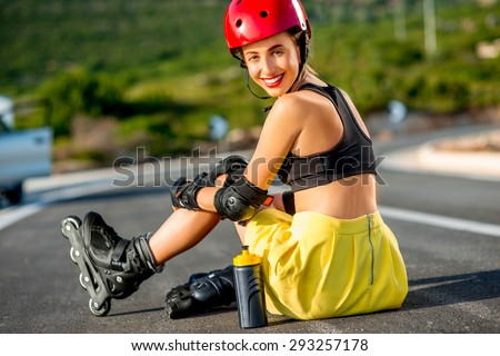 Young sport woman with rollers in yellow skirt and red helmet with full protection wear sitting on the asphalt road.