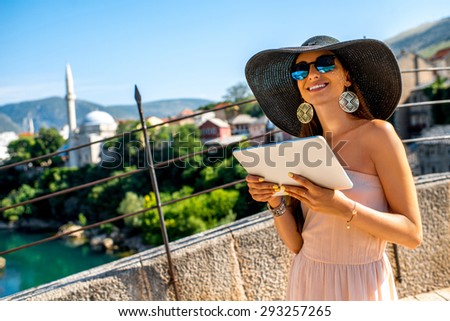 Young female tourist in dress and hat using digital tablet walking on the old bridge in Mostar city
