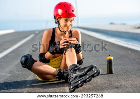 Young sport woman with rollers in yellow skirt and red helmet having knee injury sitting on the highway.