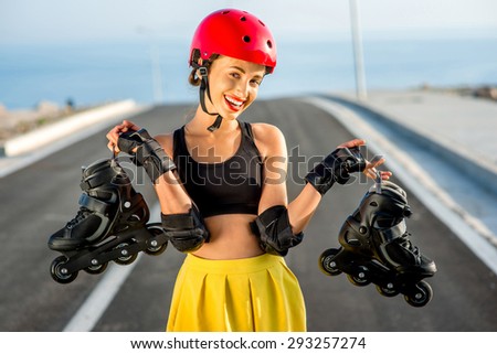 Sport woman in red helmet holding black rollers standing on the asphalt road with blue water and sky background