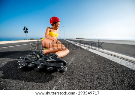 Beautiful woman in yellow swimsuit and red helmet with rollers lying on the asphalt road near the sea in summer. Wide angle view focused on rollers