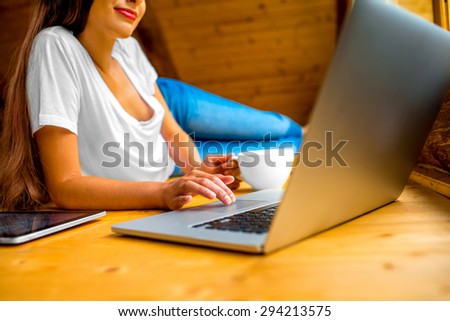 Woman in jeans and white shirt lying on the wooden floor with laptop, tablet and coffee cup in  wooden cottage. Close up view focused on hands and keyboard