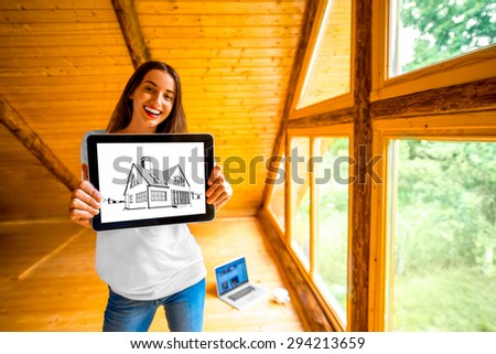 Young and cute woman showing digital tablet with house drawing near the window in cozy wooden cottage. Projecting cottage concept