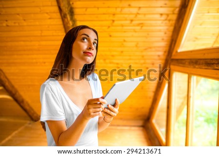 Young and cute woman thinking with digital tablet standing near the window in cozy wooden cottage. Projecting or renovating house interior concept