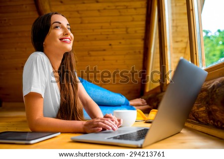 Young woman in jeans and white shirt lying on the floor with laptop, tablet and coffee cup near the window in cozy wooden cottage