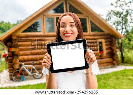 Happy woman showing digital tablet with empty screen standing near the wooden cottage.