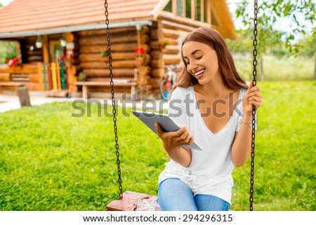 Woman sitting on a swing with digital tablet in the garden near wooden house. Eco vacations concept