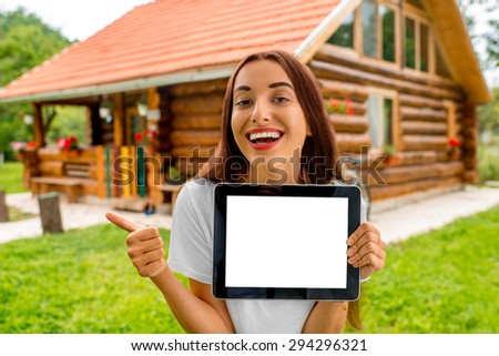 Happy woman showing digital tablet with empty screen standing near the wooden cottage.