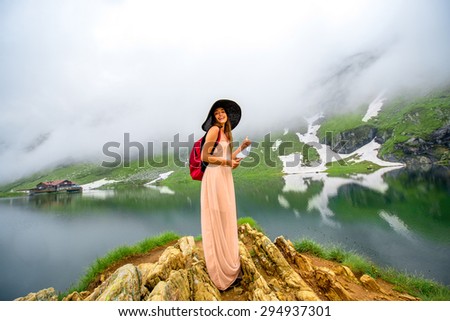 Female traveler in long dress and big hat with red backpack photographing mountain lake with fog and snow. Traveling in Romania