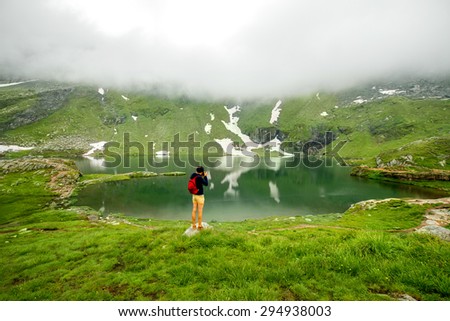 Male photographer with backpack standing on Balea lake. Traveling in Romania. Back view, general plan.
