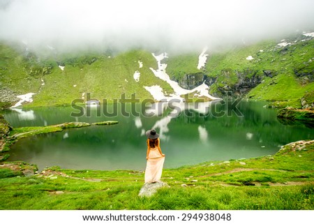 Beautiful woman in long dress and hat standing on Balea mountain lake in Romania. Back view, general plan