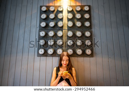 Young woman using mobile phone with many clocks on the wall background. Time phone application concept