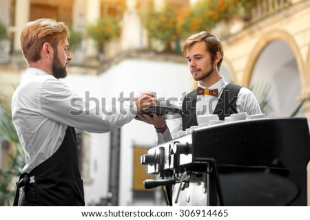 Handsome barista in uniform making coffee standing with waiter in classical renaissance cafe patio