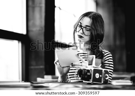 Young woman photographer looking at the printed photos with old 6x6 frame camera sitting in the loft design interior. Black and white photo