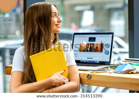 Young female student holding yellow book with laptop and colorful stuff on the table near the window with street view in the cafe or studio