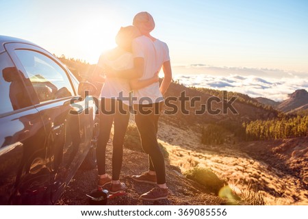 Young couple dressed alike in white t-shirt and hat embracing near the car on the roadside on the sunset.
