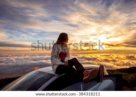 Young woman in sweater with heart shape enjoying beautiful cloudscape sitting on the car roof above the clouds on the sunrise