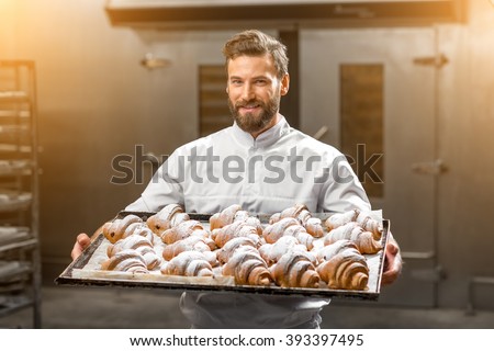 Handsome baker in uniform holding tray full of freshly baked croisants at the manufacturing