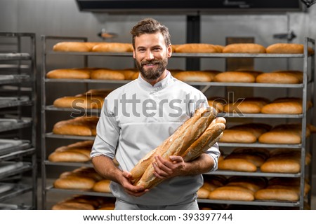 Handsome baker in uniform holding baguettes with bread shelves on the background at the manufacturing