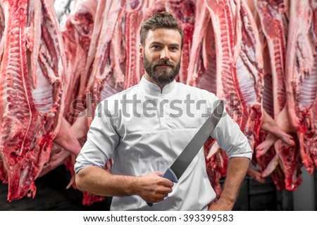 Portrait of a handsome butcher holding knife standing on the pork carcasses background at the meat manufacturing