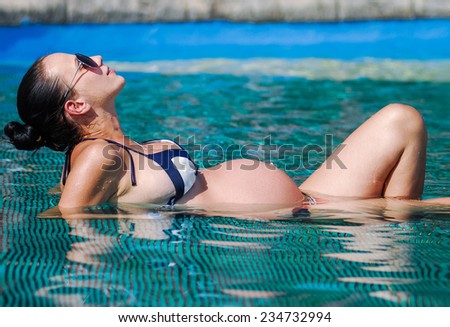 Beautiful pregnant woman sun tanning relaxed at blue pool.