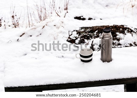 Thermos flask and coffee mug standing on a bench outdoors in snow, with out of focus background