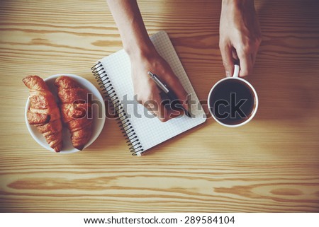 Hands with pen writing on notebook with morning coffee and croissant. View from above