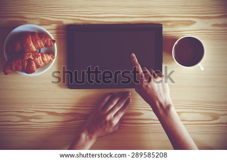 Hands holding digital tablet pc with morning coffee and croissant. View from above