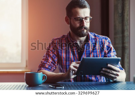 bearded man sitting with cup of morning coffee and digital tablet touching with finger
