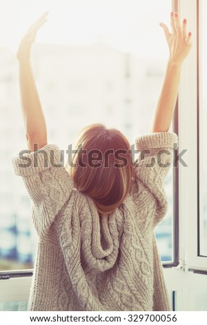 Woman near window raising hands facing the sunrise at morning