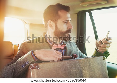 bearded businessman sitting on back seat of car with suitcase and touching phone with app