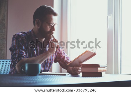 Bearded man writing with pen and reading books at table