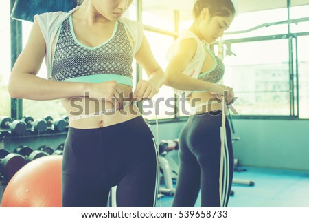 woman checking success of weight loss program with measuring tape in front of mirror at gym.