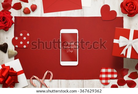 Happy Valentines day on smart phone display. Table cloth on wooden background. Love scene with hearts, gift, candle, petals, rose