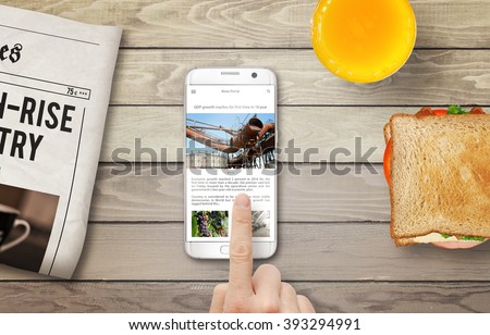 Reading news article on the phone during breakfast. Newspapers, juice and sandwich on the table with top view.