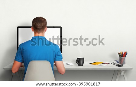 Man working on computer with isolated screen in office interior. Work desk with keyboard, mouse, cup of coffee, paper, pencils. Free space on wall for text.