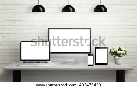 Devices on desk with isolated screen for mockup. Computer display, laptop, tablet and smart phone on office desk. Flowers, lamps and brick wall in background.
