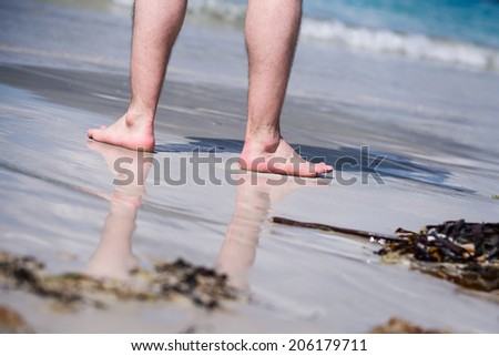 Male bare feet in a warm sand, man taking a walk on a sunny beach with turquoise water during vacation.