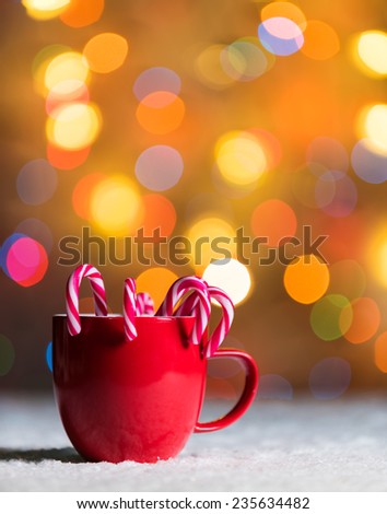 Red cup, red mug with candy canes in snow with defocussed fairy lights, bokeh in the background, Festive Christmas background with copyspace