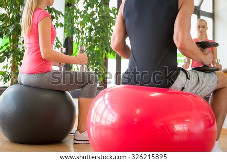 Young couple training in gym with dumbbells in front of a mirror sitting on fitballs