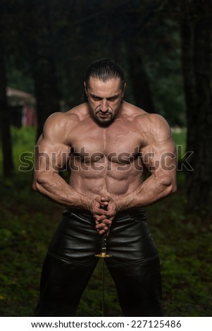 Dangerous Man Portrait With Ancient Sword - Standing In Forest Wearing Leather Pants