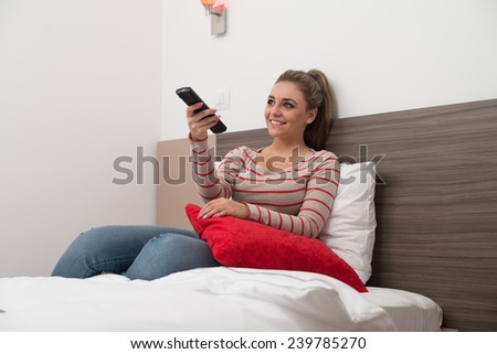 Beautiful Young Woman Watching Television Sitting On Bed With The Remote Control In Her Hand