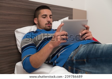 Young Male Student Lying On Bed And Having Fun With Touch Pad In Bedroom