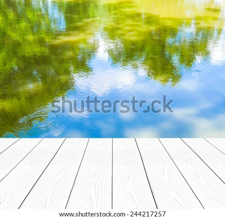 Empty perspective white wood over the reflection of green trees and blue sky on water, product display template