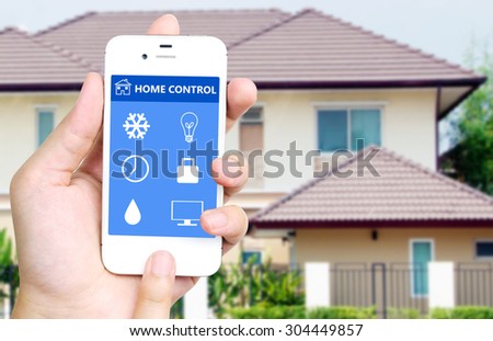 Hand holding white mobile smart phone with smart home application on the screen over blurred house background, smart home concept
