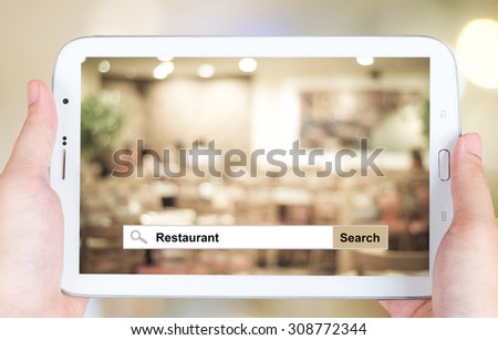 Hand holding tablet with restaurant word on search bar over blurred restaurant background, restaurant reservation, food online, food delivery concept